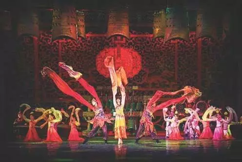 Group of dancers performing on stage with red and pink costumes and large flowing fabric props, with ornate backdrop and lighting.