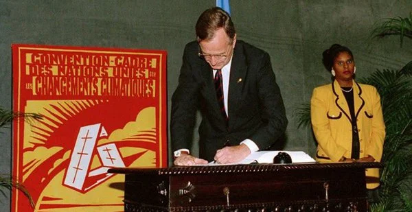 A man signing a document at a table, with a woman standing nearby, in front of a red and yellow poster that reads 'CONVENTION-CADRE DES NATIONS UNIES CHANGEMENT CLIMATIQUE'.