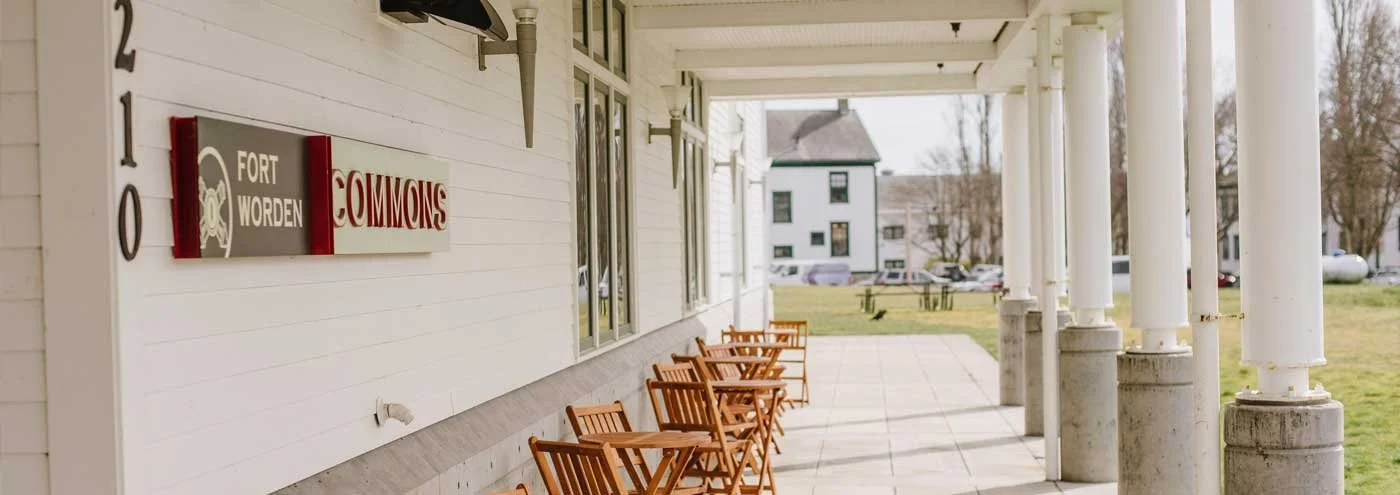 A covered porch with several wooden chairs lined up against a white building, with a sign that reads 'Fort Worden Commons' on the wall. The background shows a grassy area, trees, and some buildings.