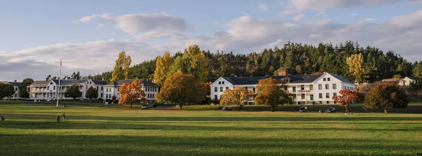 Residential buildings with white walls and dark roofs, surrounded by trees with autumn foliage, on a hillside overlooking a large grassy park with people walking and cars parked along the street.