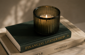 A lit candle in a dark glass holder placed on top of a thick financial leadership book, with a second book titled 'TENOCLÉ' underneath, on a light-colored surface.