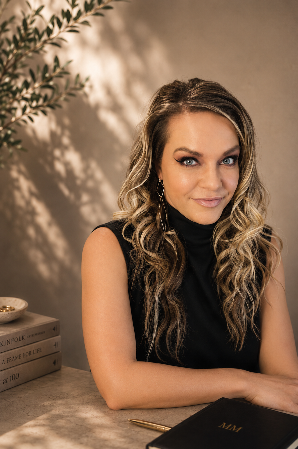 A woman with wavy blonde hair and blue eyes wearing a black sleeveless turtleneck, sitting at a desk with a black planner, a gold pen, and a stack of books, with a plant and shadowed background.