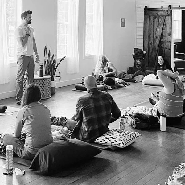 A group of people participating in a meditation or yoga session in a cozy, well-lit indoor space, sitting on cushions on the wooden floor, with a standing instructor guiding them.