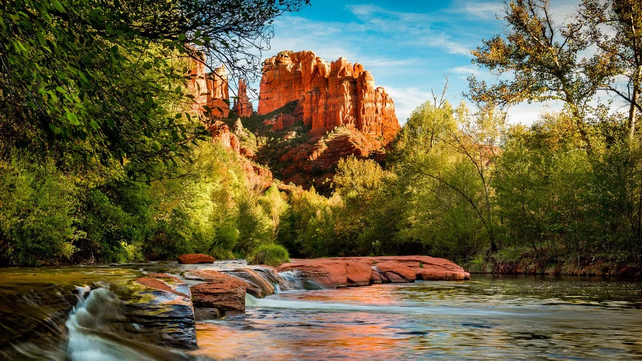 A landscape of a flowing river with rocks, green trees, and red rock formations in the distance under a blue sky.