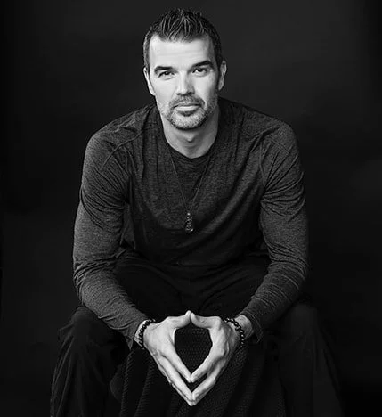 A man with dark hair and a beard sitting against a black background, wearing a long-sleeve dark shirt and making a heart shape with his hands.