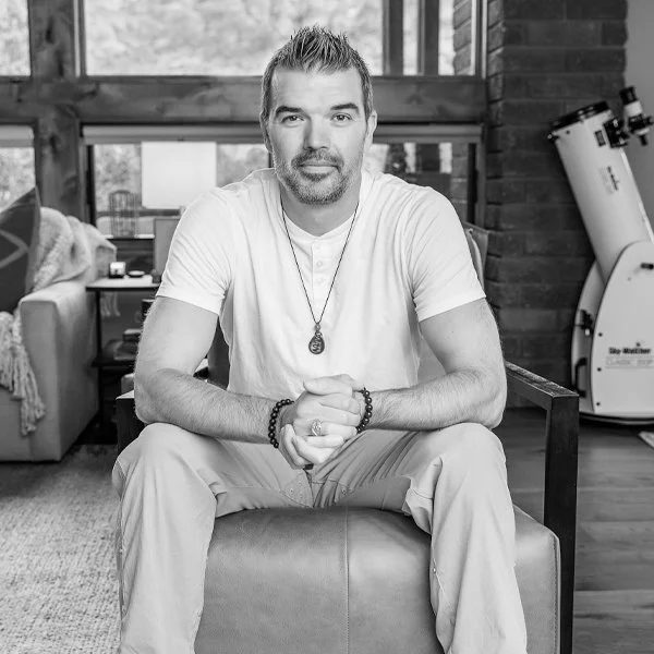A man with short hair and a beard sits on an ottoman in a living room with wooden walls and a brick corner. He's wearing a white t-shirt, beaded bracelets, a necklace, and khaki pants. He has his hands clasped and is looking at the camera.