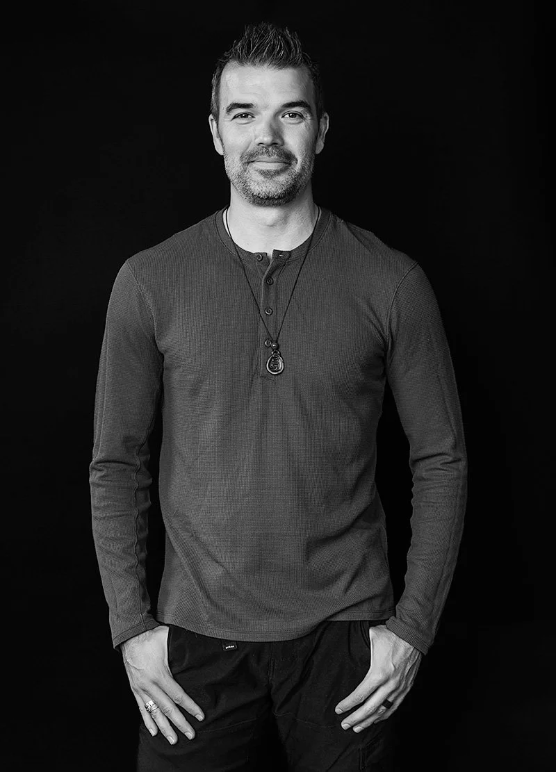 Black and white photo of a man with short hair and a beard, wearing a long-sleeve shirt and a necklace, standing against a dark background.