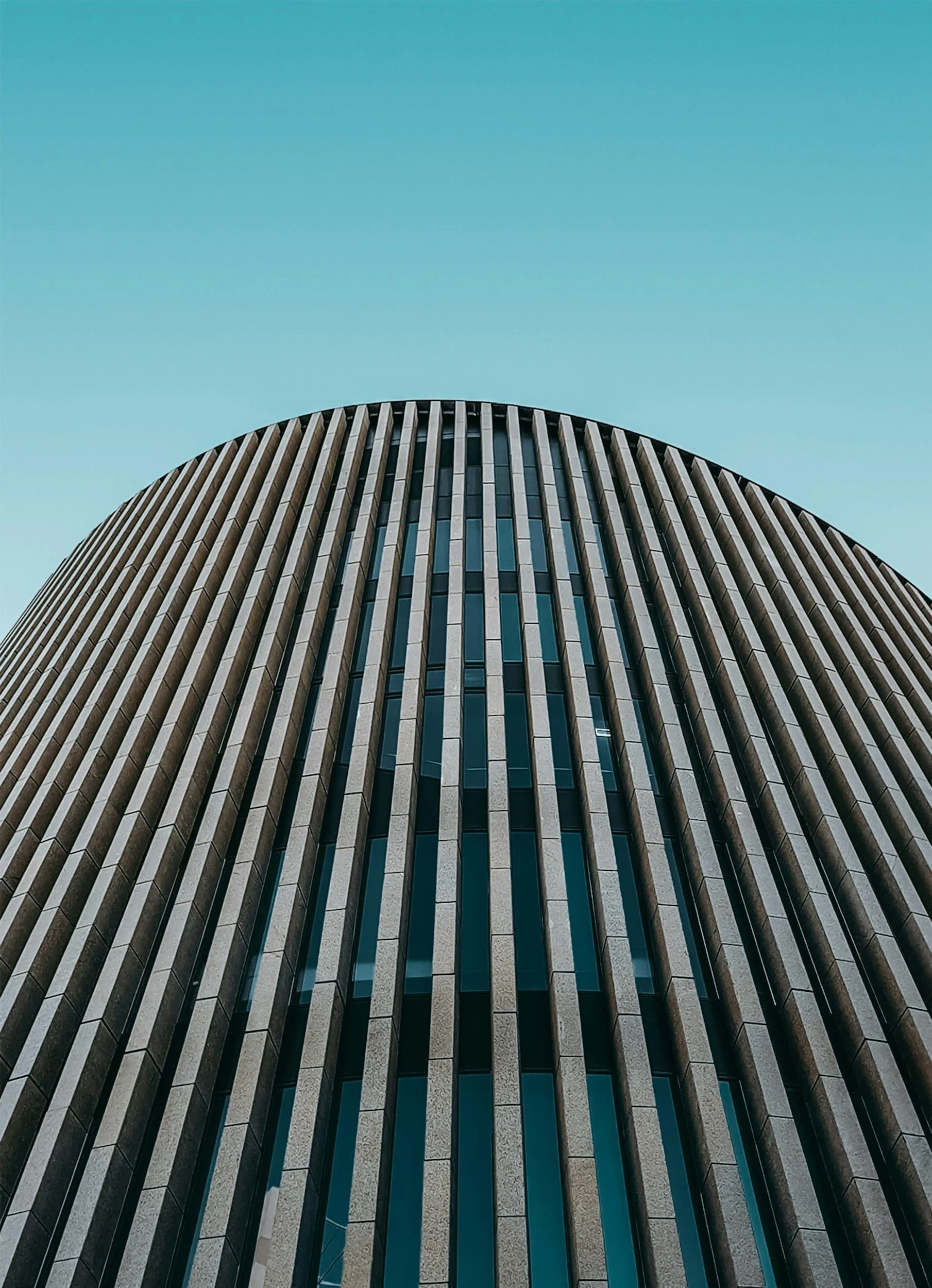 Low-angle view of a modern, curved high-rise building with vertical beige and black lines, set against a clear blue sky.