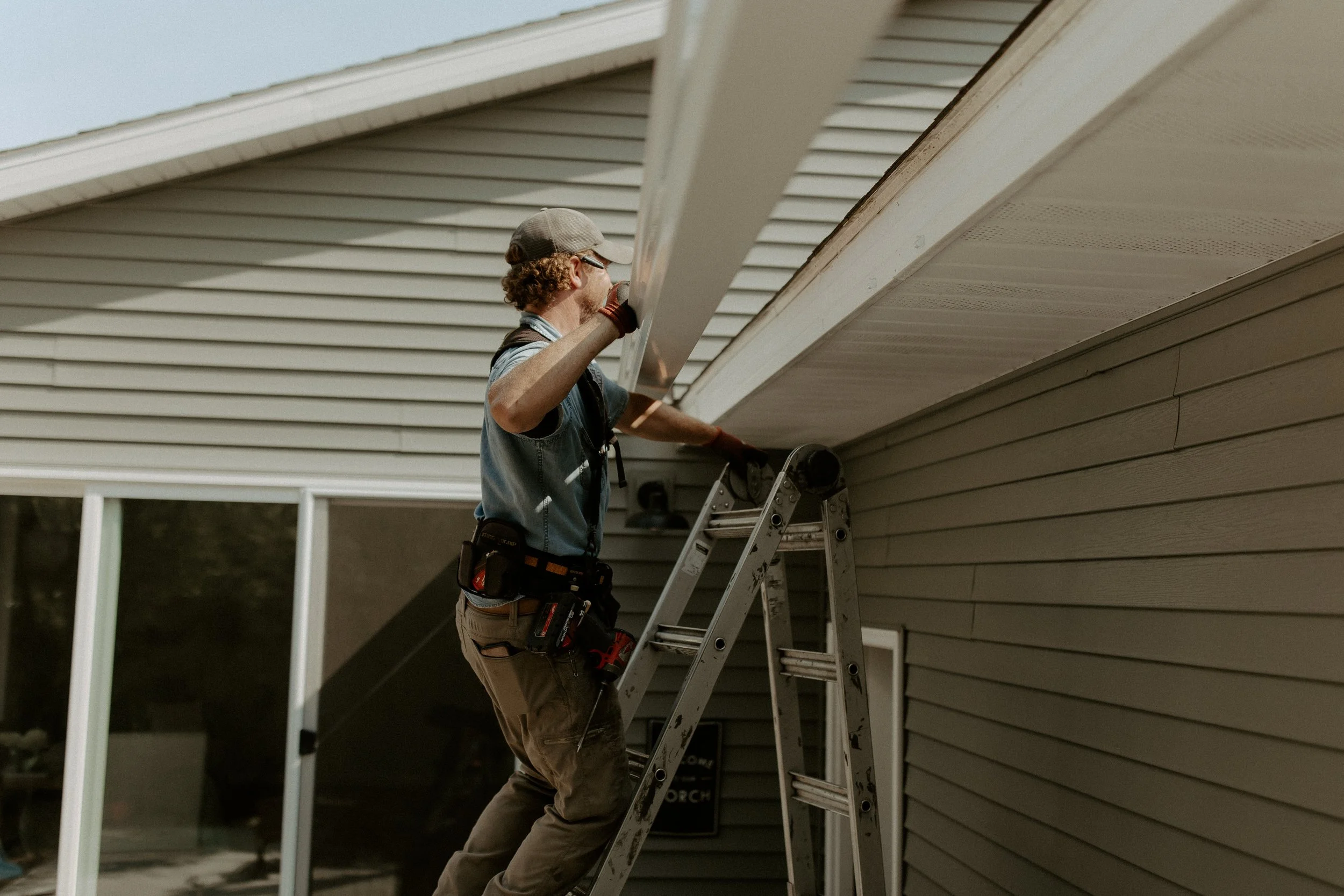 A man wearing a hat stands on a ladder installing gutters on a house with beige siding.