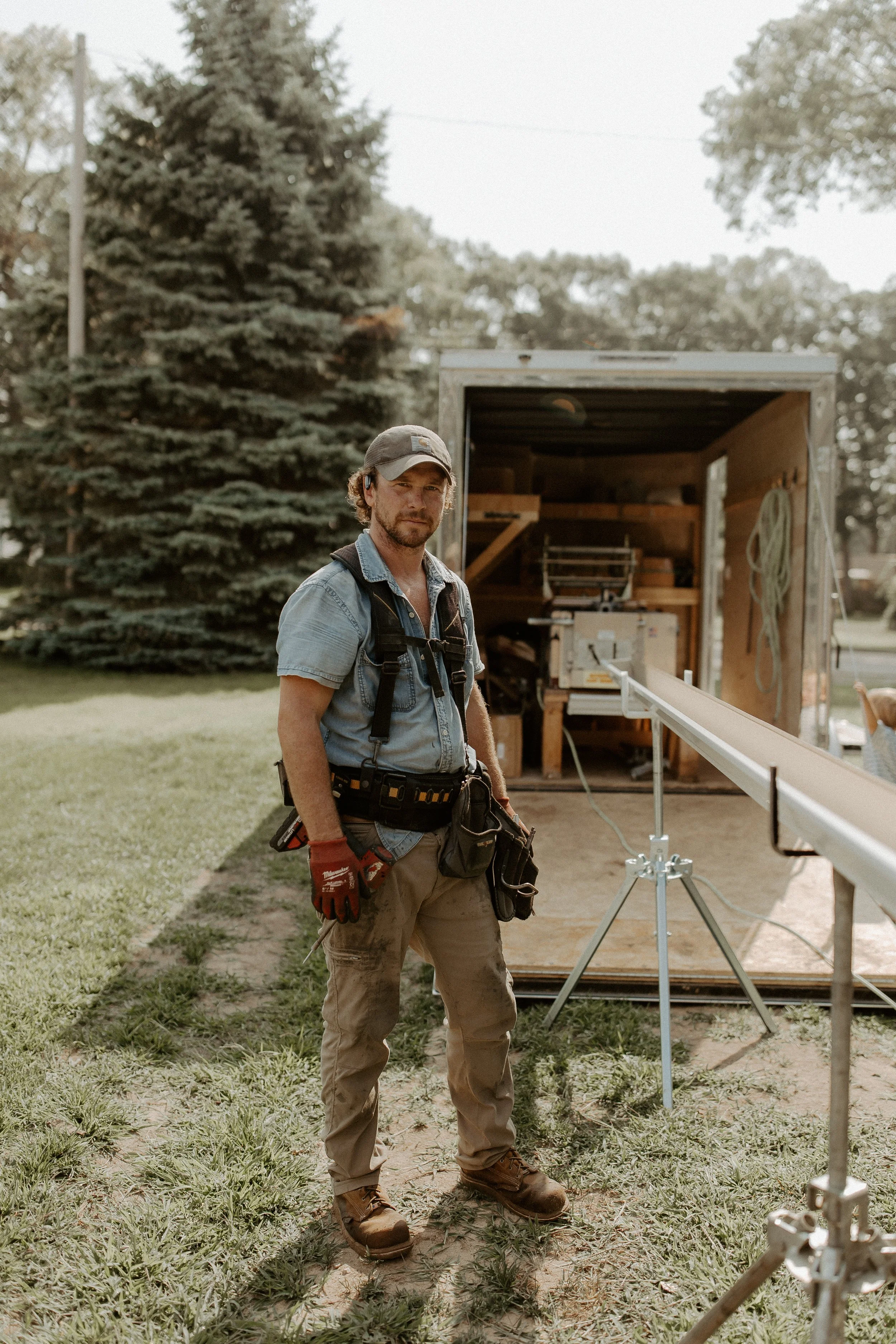 A man in work clothes, wearing a hat and red gloves, stands outdoors in front of a trailer with woodworking equipment inside. He is near a construction setup with a level and other tools, and there are trees in the background.