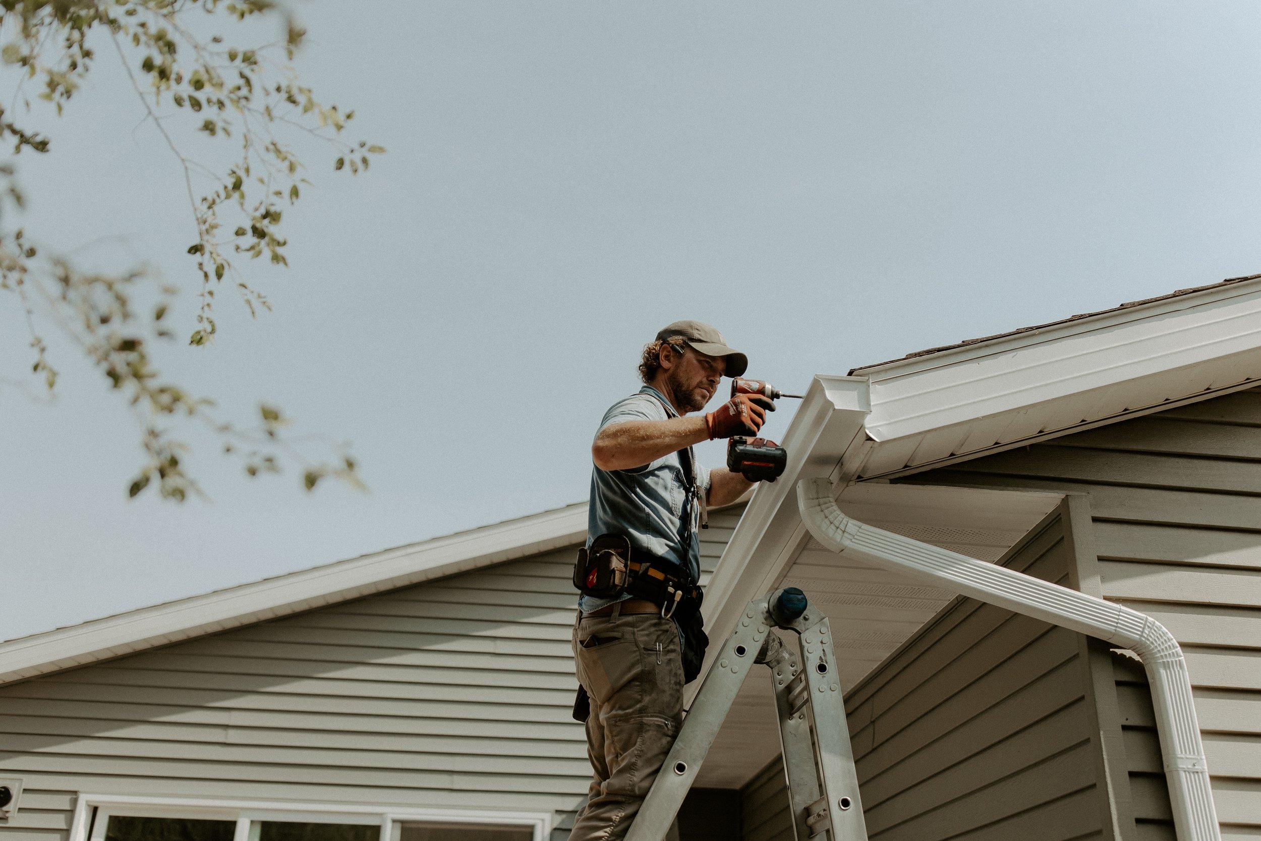 A man standing on a ladder installing new gutters and downspouts on the roof of a house in Muskegon County, using a cordless drill, wearing a cap, gloves, and a tool belt, with a light blue sky in the background.