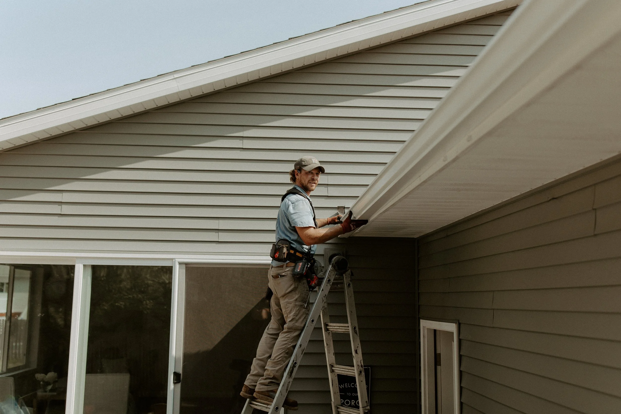 A man standing on a ladder installing or repairing the soffit under the roof overhang of a house with gray siding and large windows.