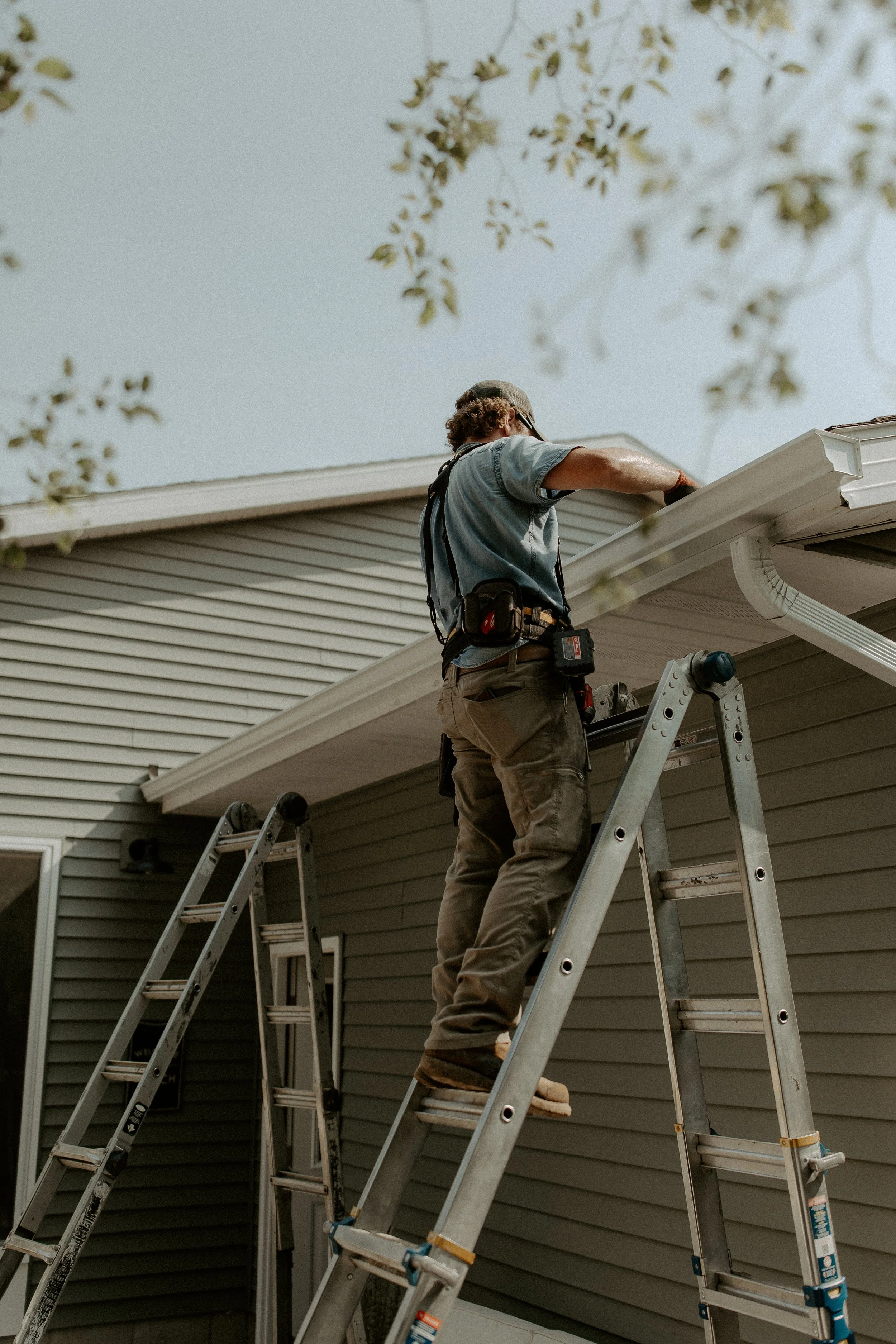 A worker standing on a stepladder installing gutter on a house. The house has beige siding, and the worker is wearing a gray shirt, khaki pants, and a gray cap.