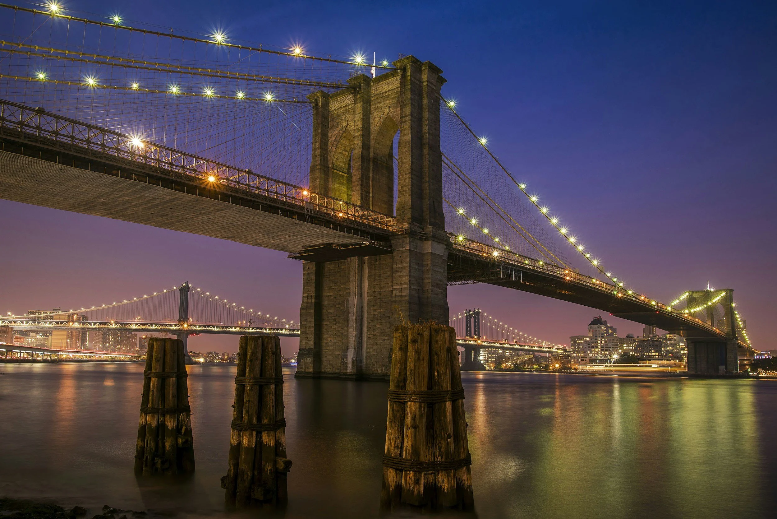 The Brooklyn Bridge illuminated at night with city lights and Hudson River reflection.