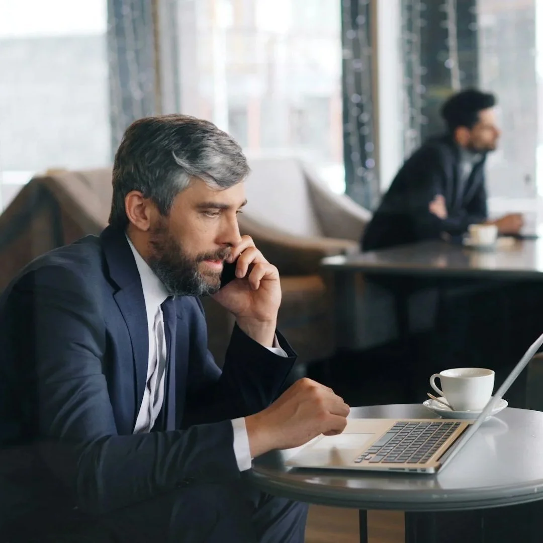 A middle-aged man in a business suit using a laptop in a cafe, with a cup of coffee on the table, and another man in the background sitting at a table.