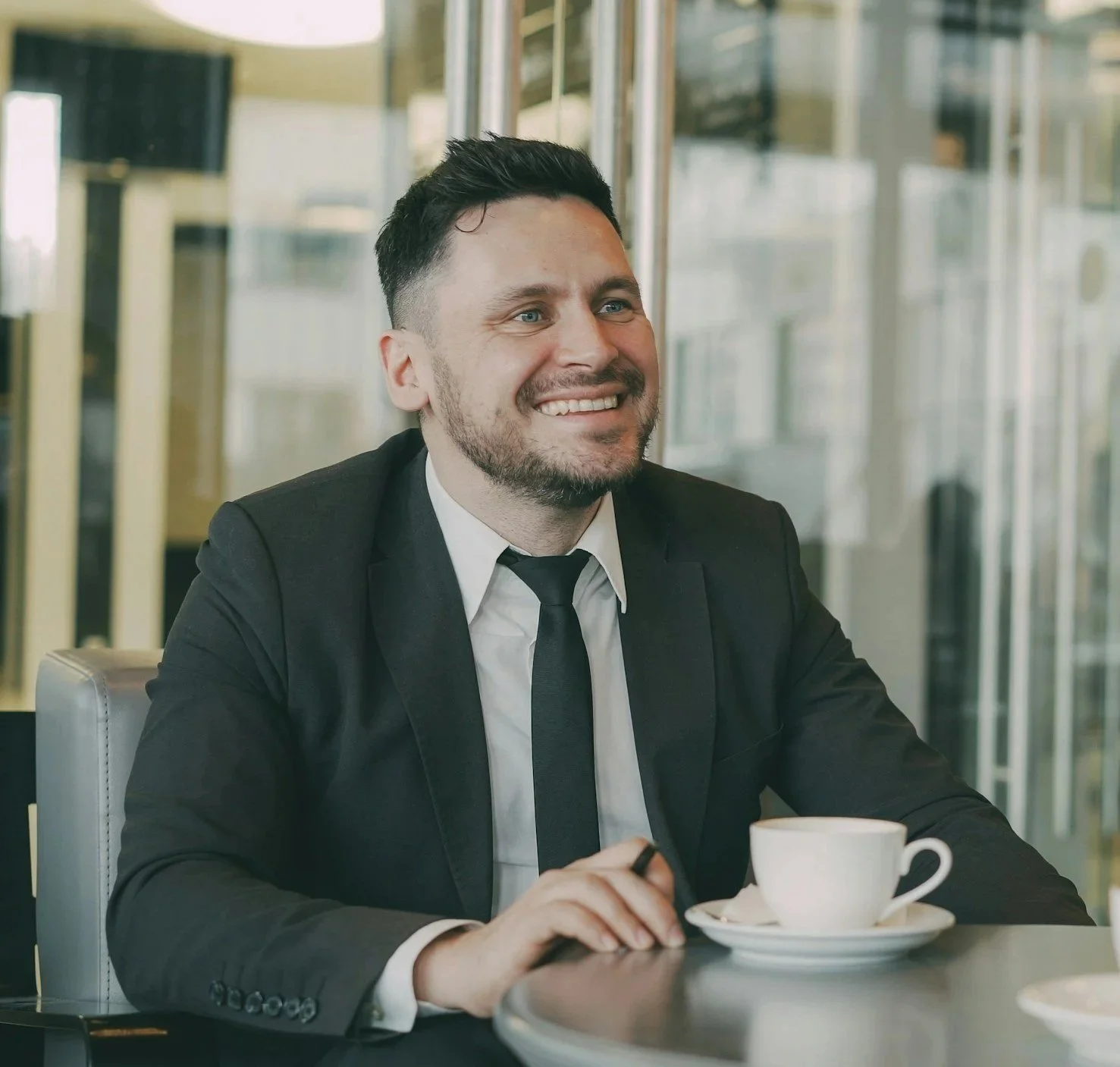 A smiling man in a black suit, white shirt, and black tie sitting at a table with a white coffee cup and saucer, in a bright office or conference room with glass walls.