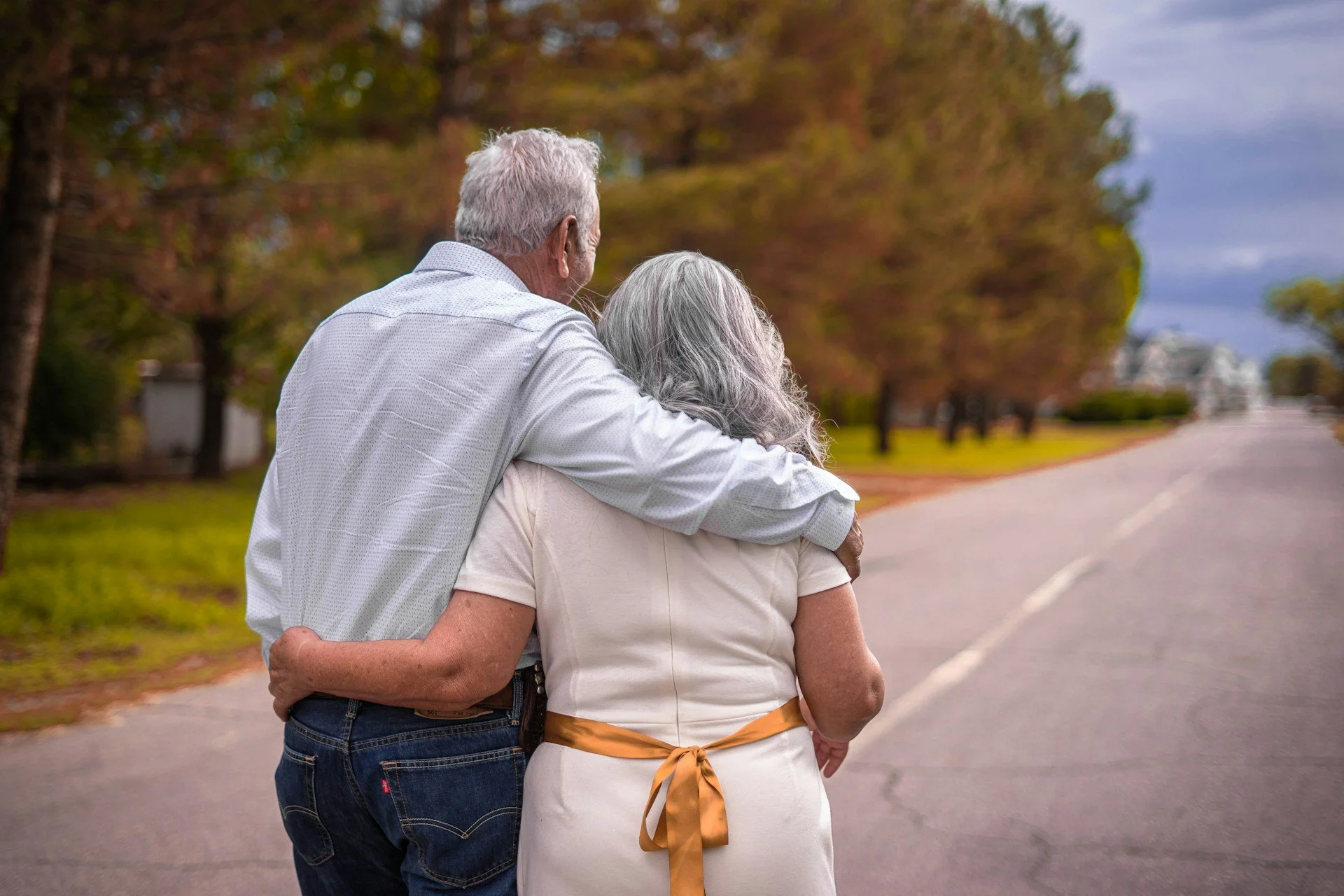 An elderly couple walking together outdoors on a street lined with trees during fall, with the man hugging the woman from behind.