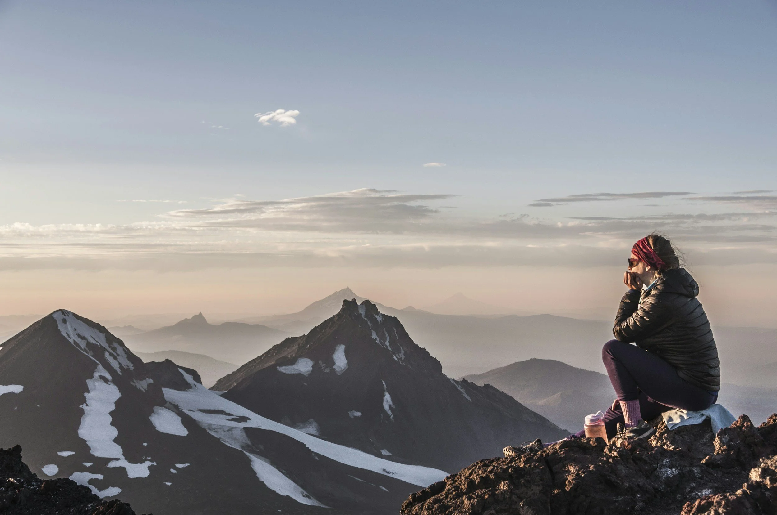 A woman sitting on a mountain ridge, gazing at snow-capped peaks and distant mountains during sunset or sunrise in a rugged landscape.
