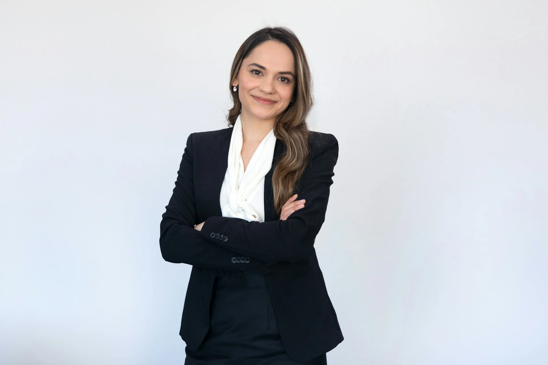 A professional woman with long brown hair, smiling, wearing a black blazer and a white blouse, standing against a plain white background.