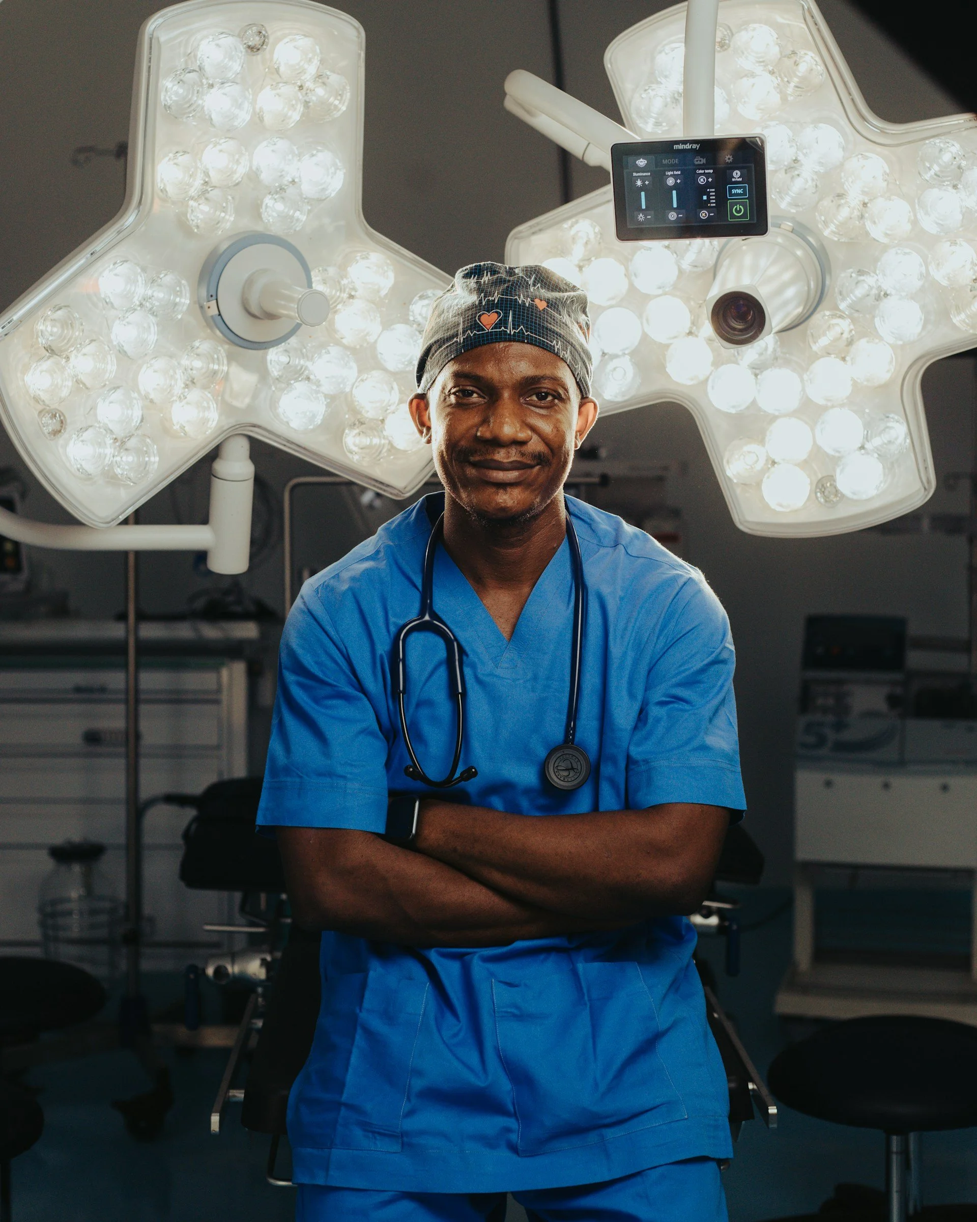 A male surgeon wearing blue scrubs and a patterned surgical cap with hearts and heartbeat line, standing in an operating room with bright surgical lights and medical equipment in the background.