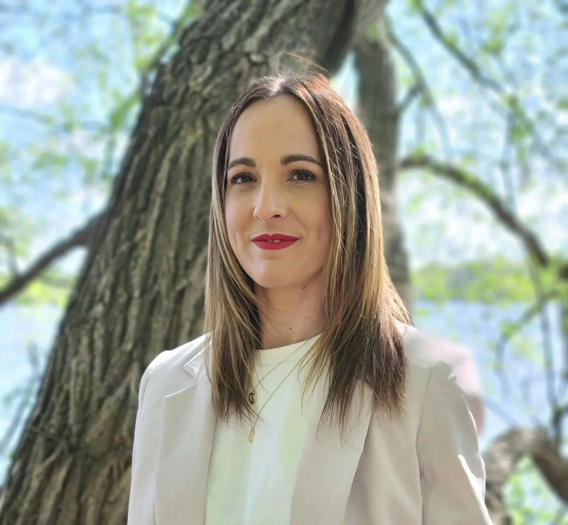 Close-up of a smiling woman with long brown hair and red lipstick standing outdoors near a tree with green leaves.