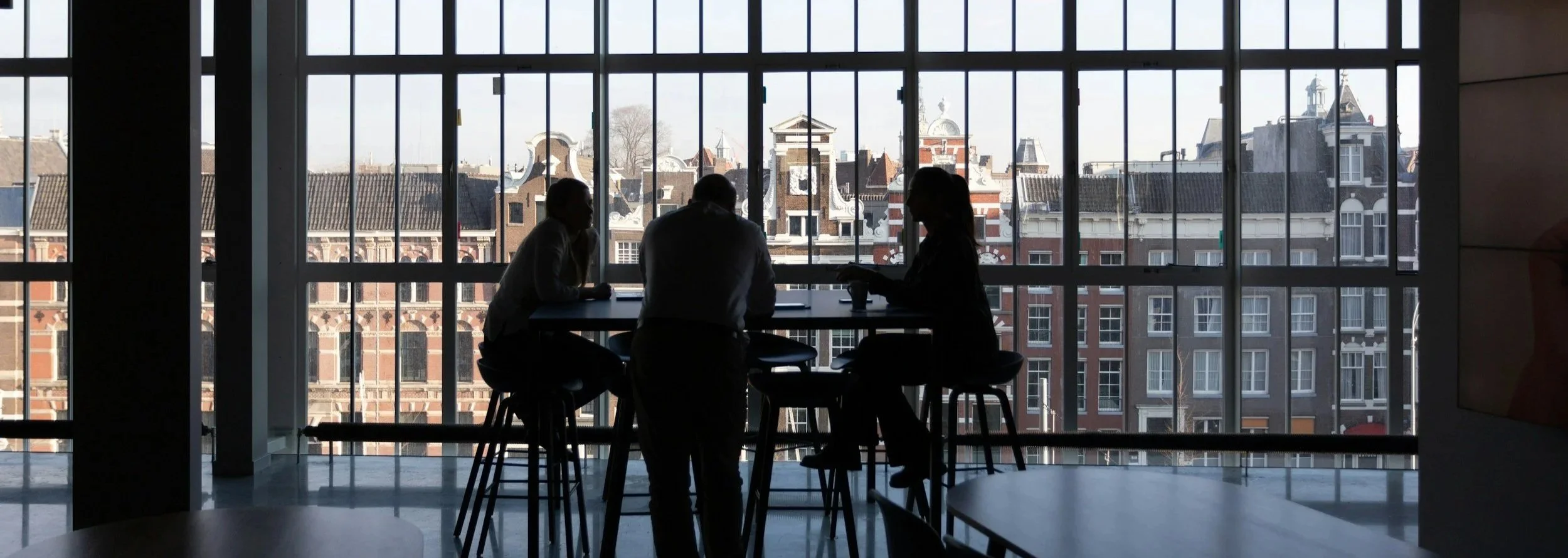 Silhouettes of three people sitting and standing at a high table in front of large window showing city buildings outside, some with ornate rooftops and styles.