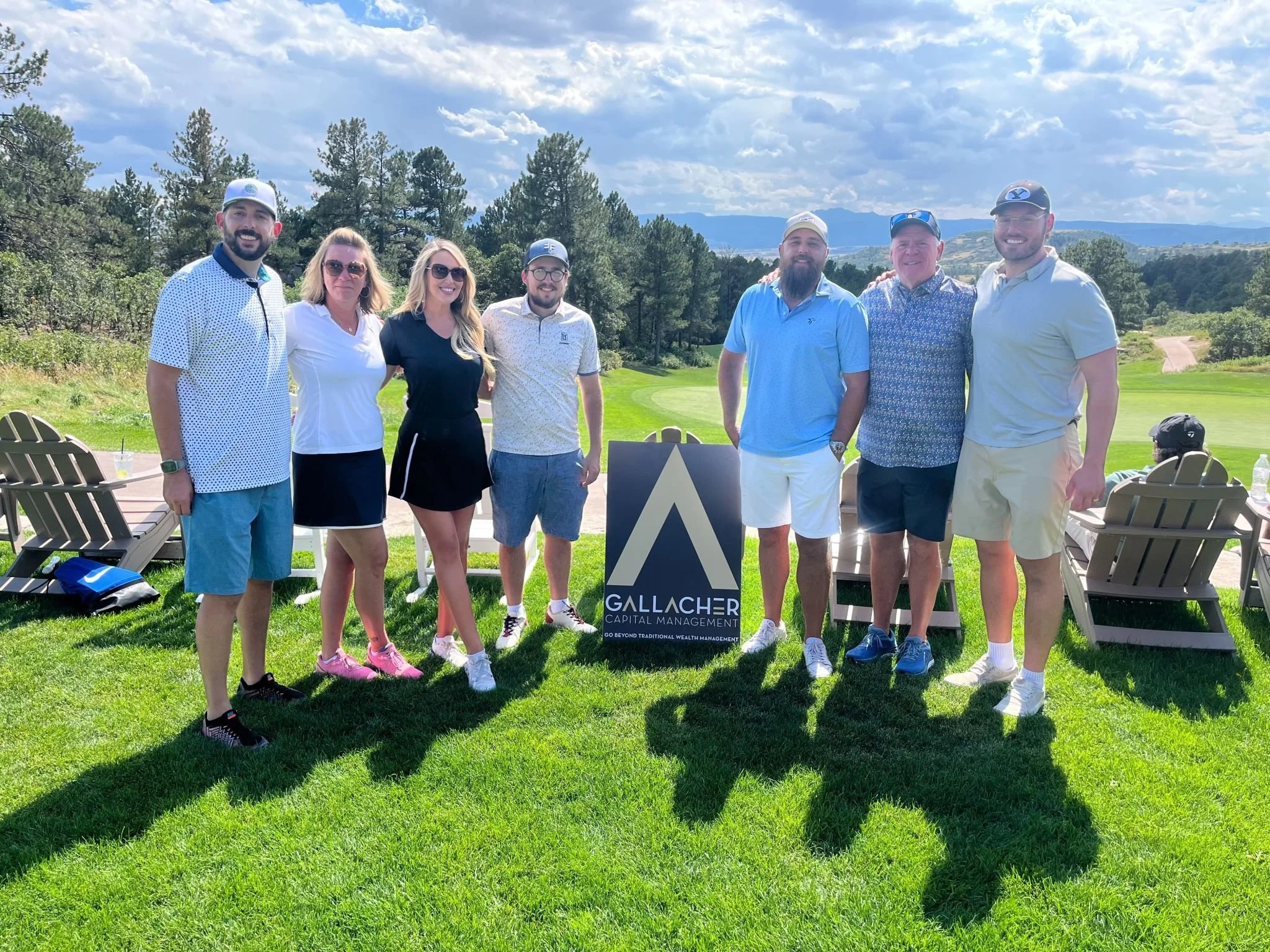 A group of seven people standing outdoors on a golf course, smiling, with a sign for Gallacher Capital Management and scenic mountains and trees in the background.