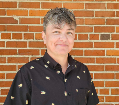 Person with short gray curly hair smiling in front of a brick wall, wearing a black shirt with small colorful patterns.