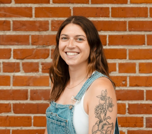Young woman with brown hair smiling, wearing a white shirt and denim overalls, standing in front of a brick wall.