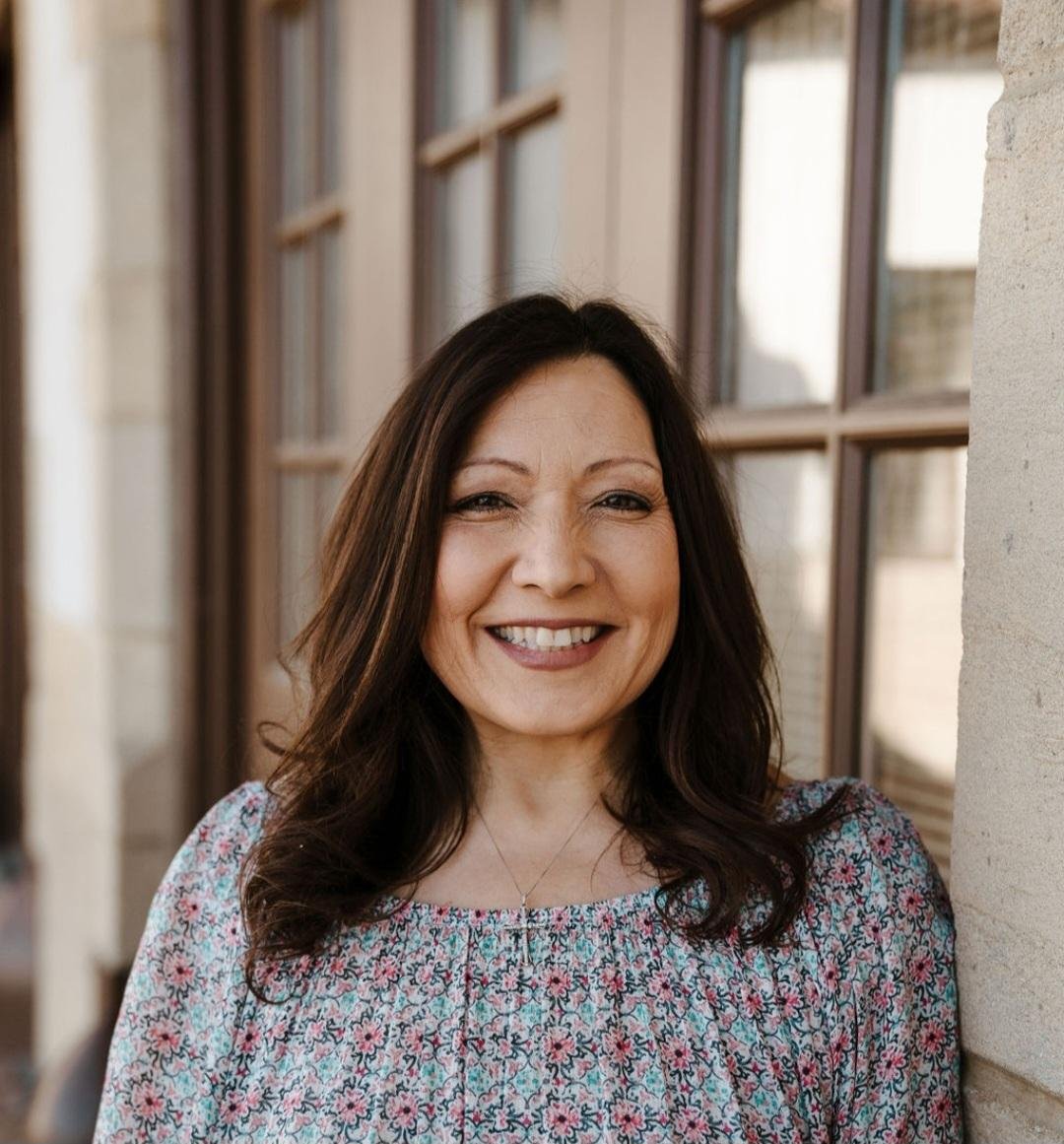 A woman with dark brown hair smiling outdoors, standing beside a beige wall and a large window with multiple panes during daytime.