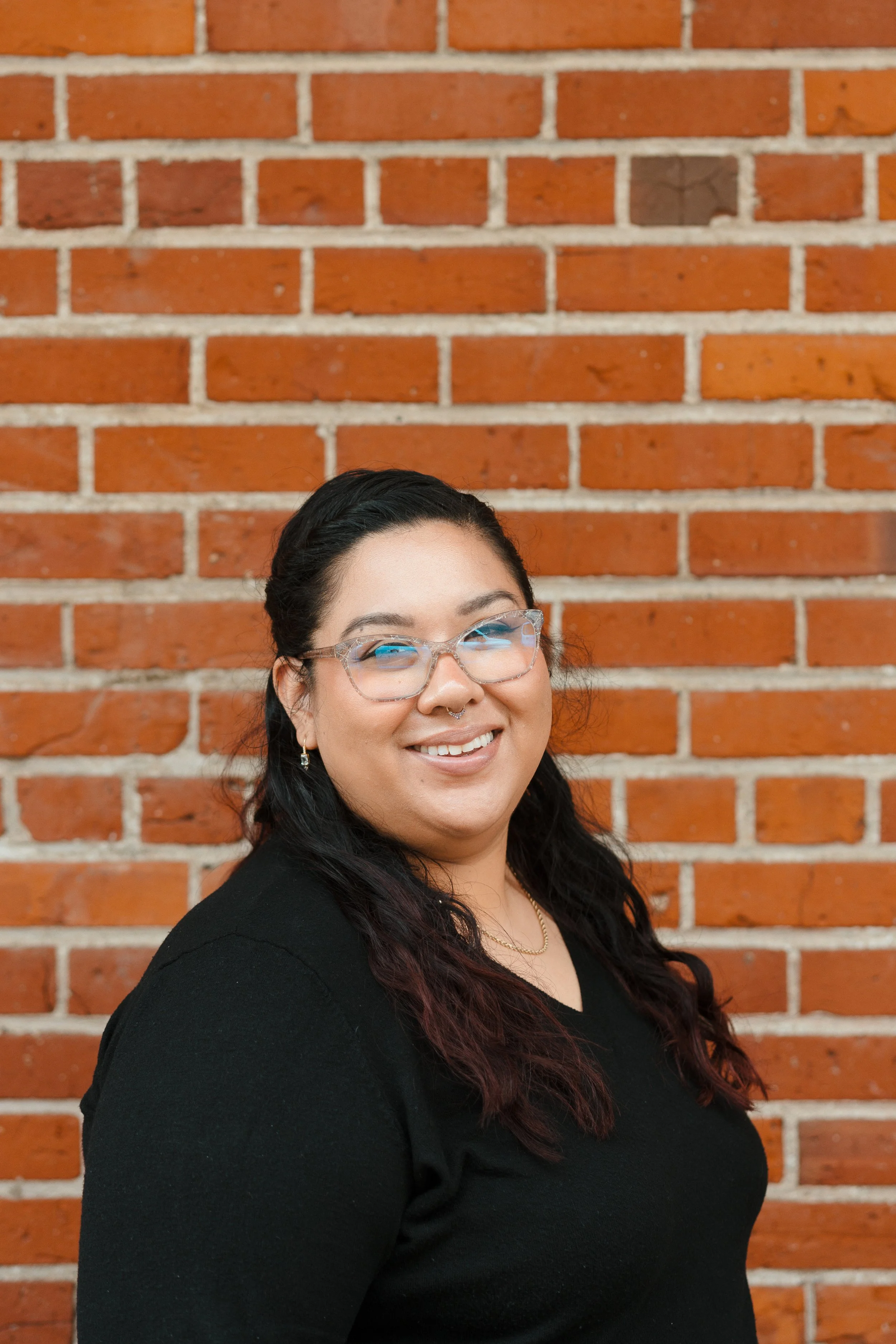 A woman with black hair and glasses smiling in front of a brick wall