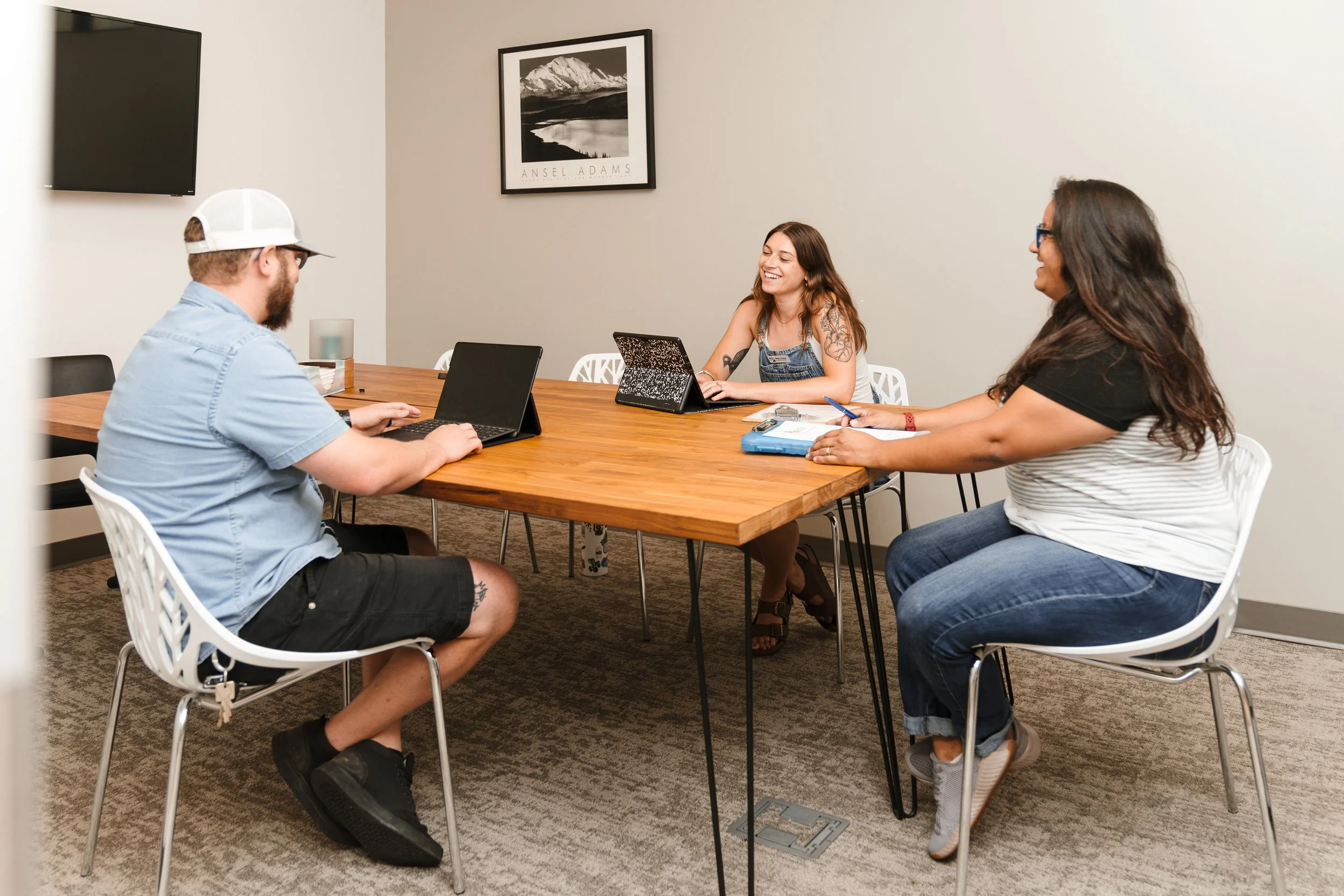 Four people sitting around a wooden table in a meeting room, engaging in conversation and smiling, with a framed black-and-white mountain photograph on the wall in the background.