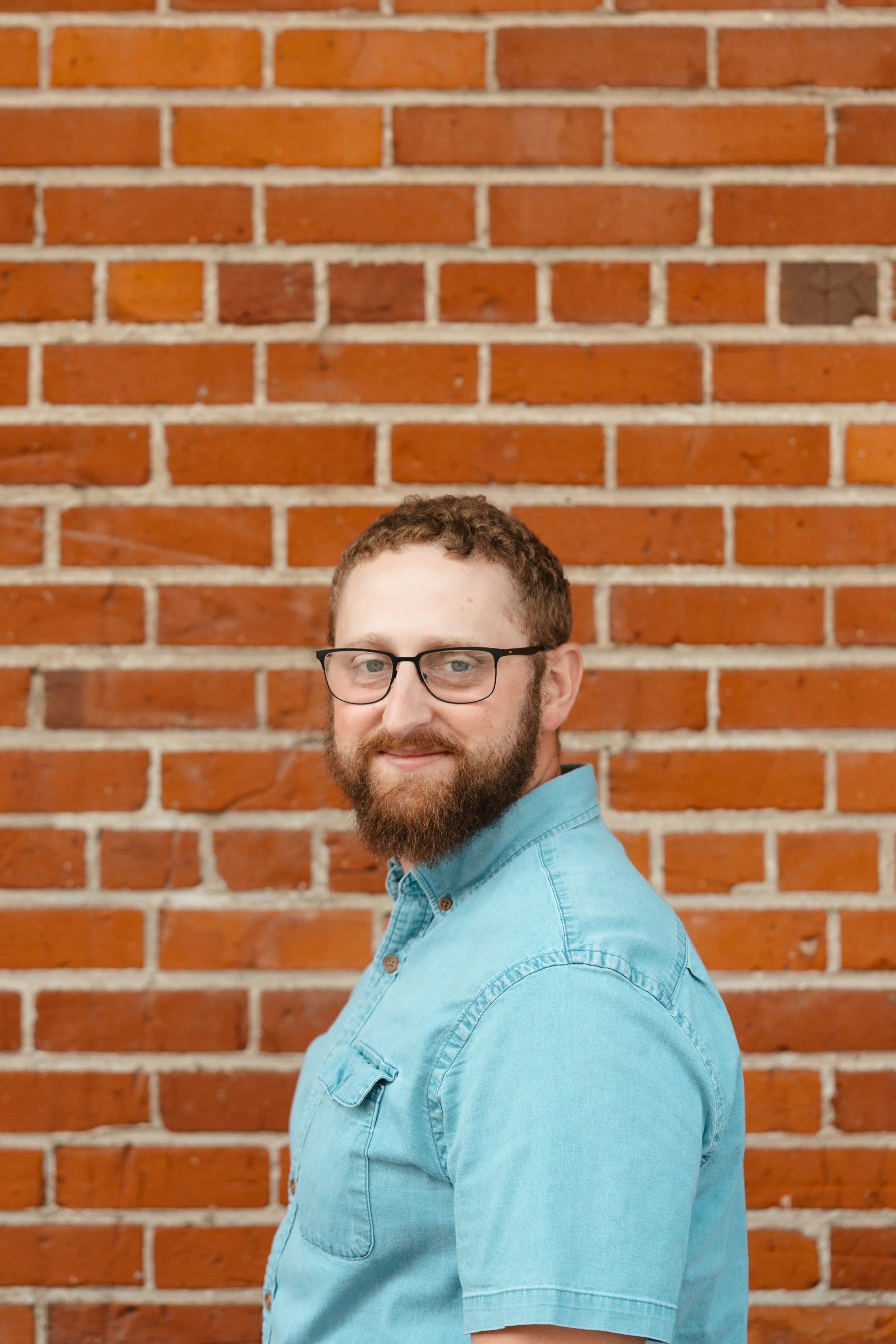 A man with glasses and a beard wearing a light blue denim shirt, standing in front of a brick wall.