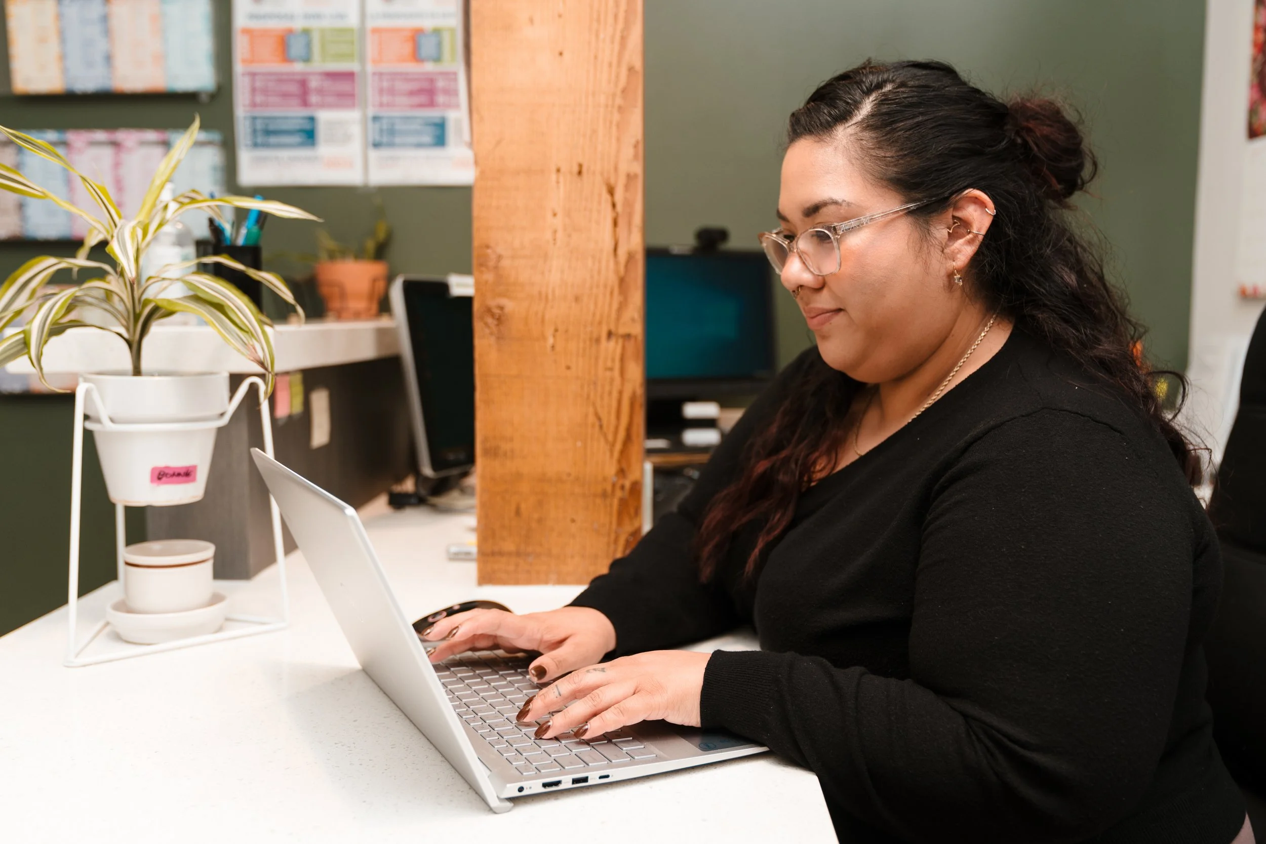 A woman with dark hair and glasses working on a laptop at a white desk in an office space, with a green wall, a wooden partition, a potted plant, and colorful posters on the wall in the background.