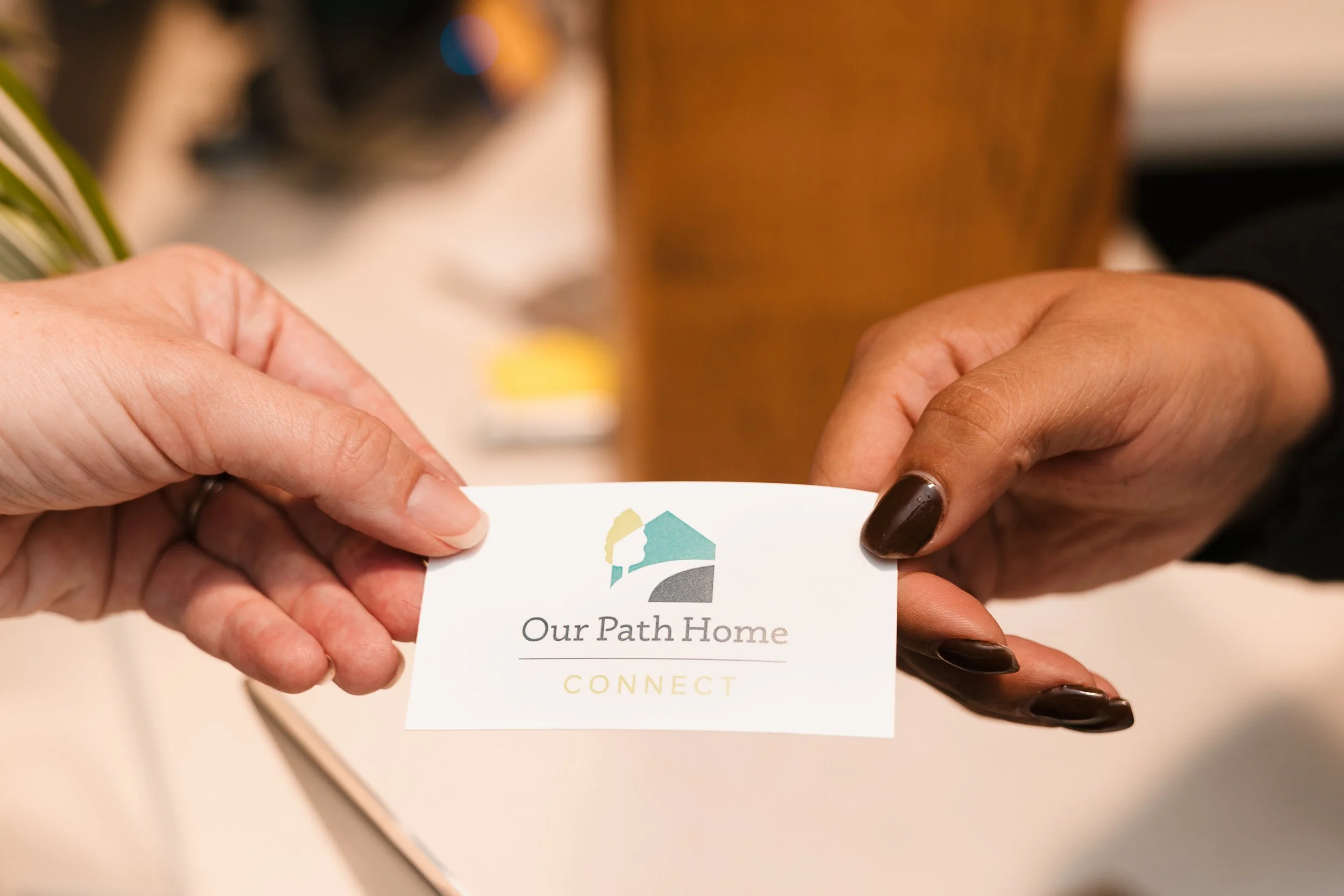 Close-up of hands holding a business card with the logo and text "Our Path Home CONNECT".