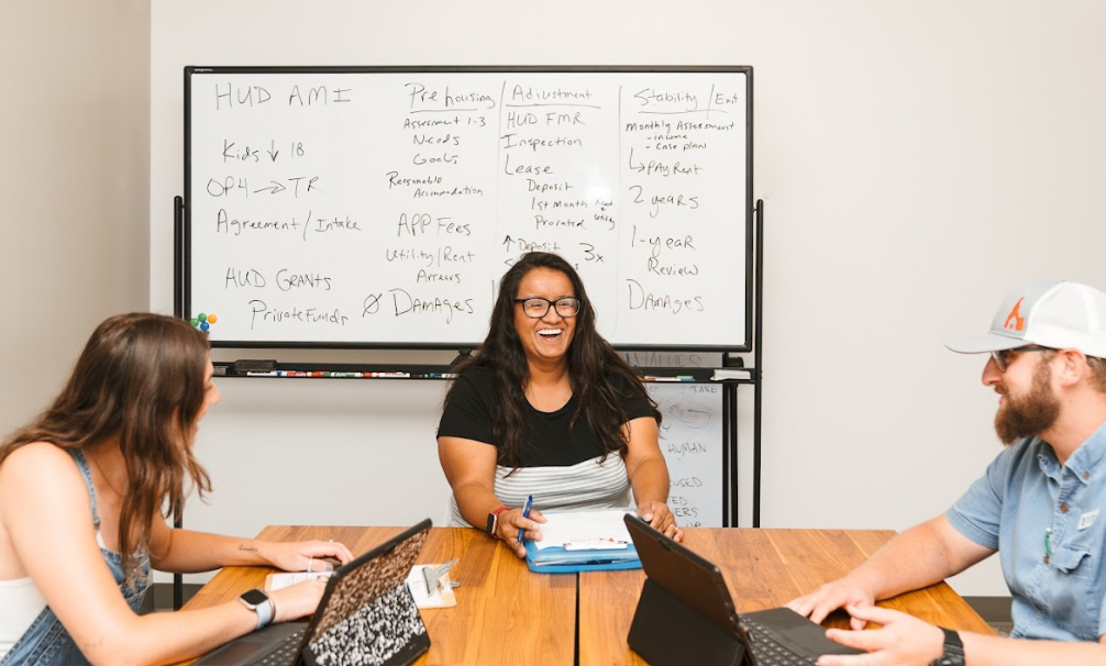 Three people sitting at a wooden table in a meeting room, with a whiteboard filled with notes behind them. The woman in the middle is laughing and holding a pen, while the woman on the left and the man on the right are engaged in the discussion.