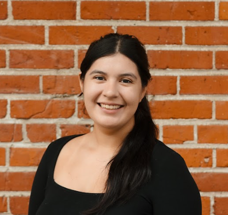 Portrait of a young woman with long dark hair, smiling, wearing a black top, standing against a red brick wall.