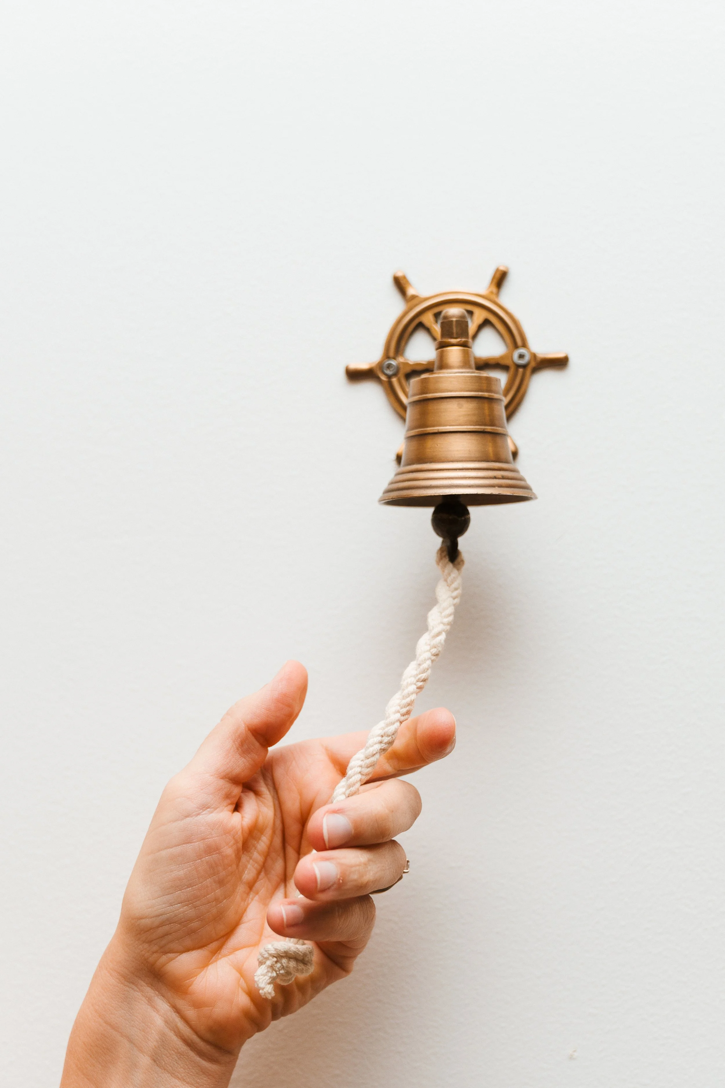 A person holding a small bell made of metal, attached to a white rope, against a plain white background.