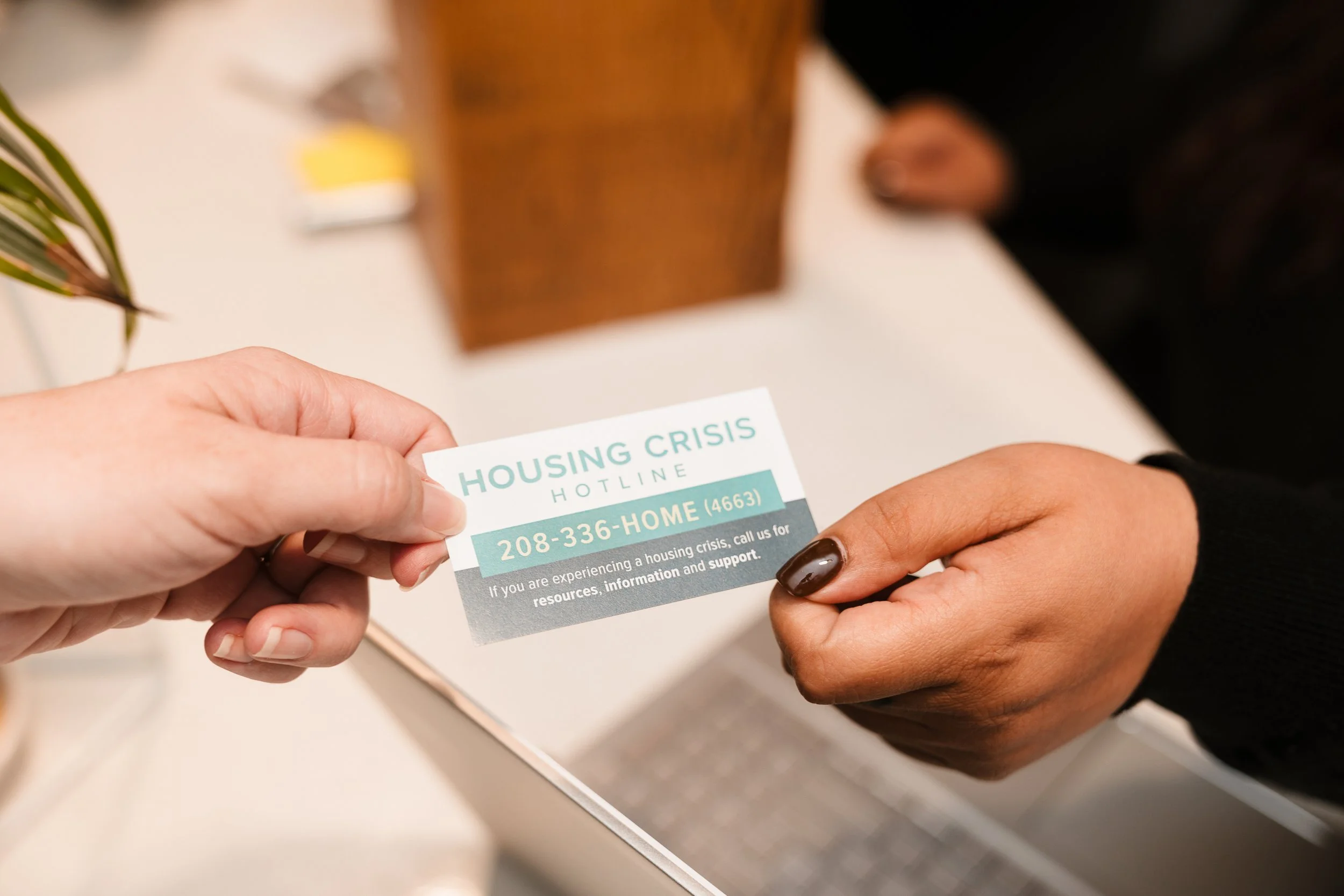 Close-up of two hands exchanging a flyer about housing crisis hotline, with a blurred indoor background.
