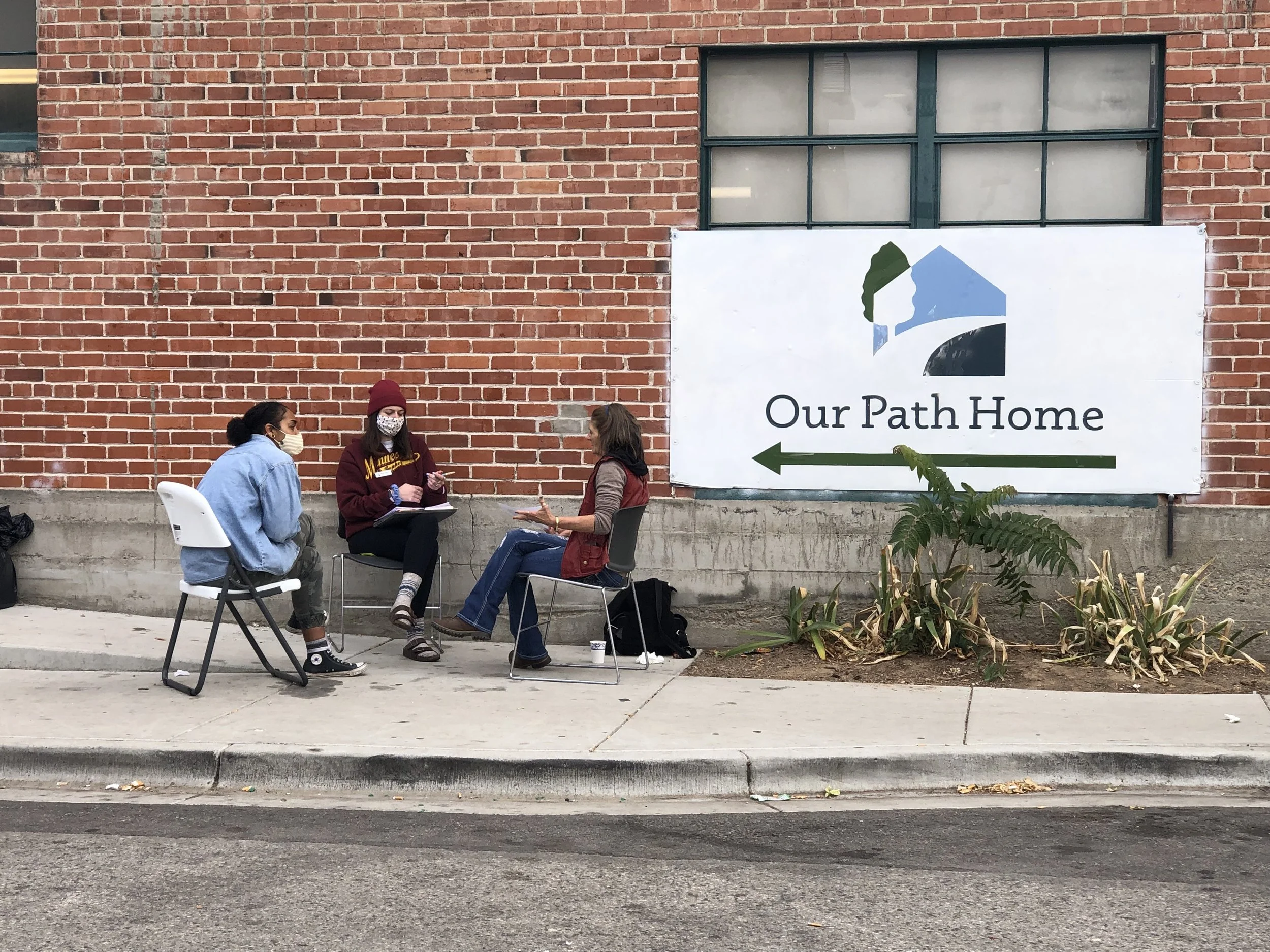 Three women wearing masks sitting and talking outside in front of a brick building with a sign that says 'Our Path Home' and an arrow pointing left.