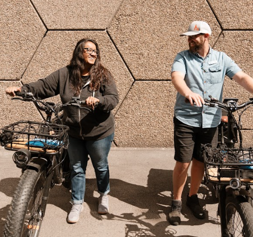 A woman and a man standing next to electric bikes outdoors in front of a textured wall, smiling and talking.