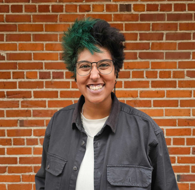 A smiling woman with short, curly hair and glasses, standing in front of a brick wall.