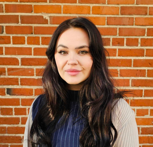 portrait of a woman with long dark hair standing in front of a brick wall