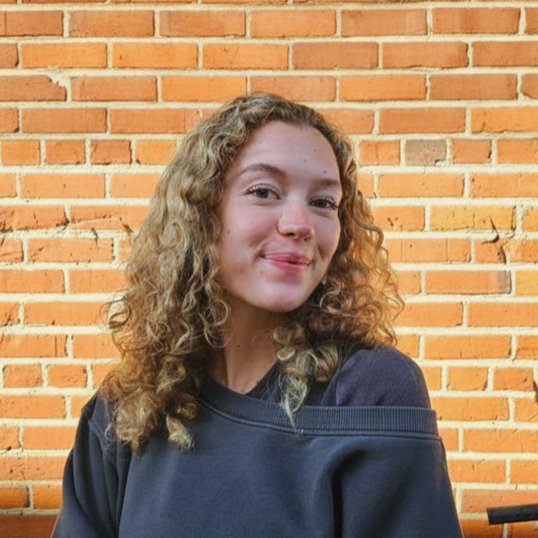 A smiling young woman with curly blonde hair standing against a brick wall.
