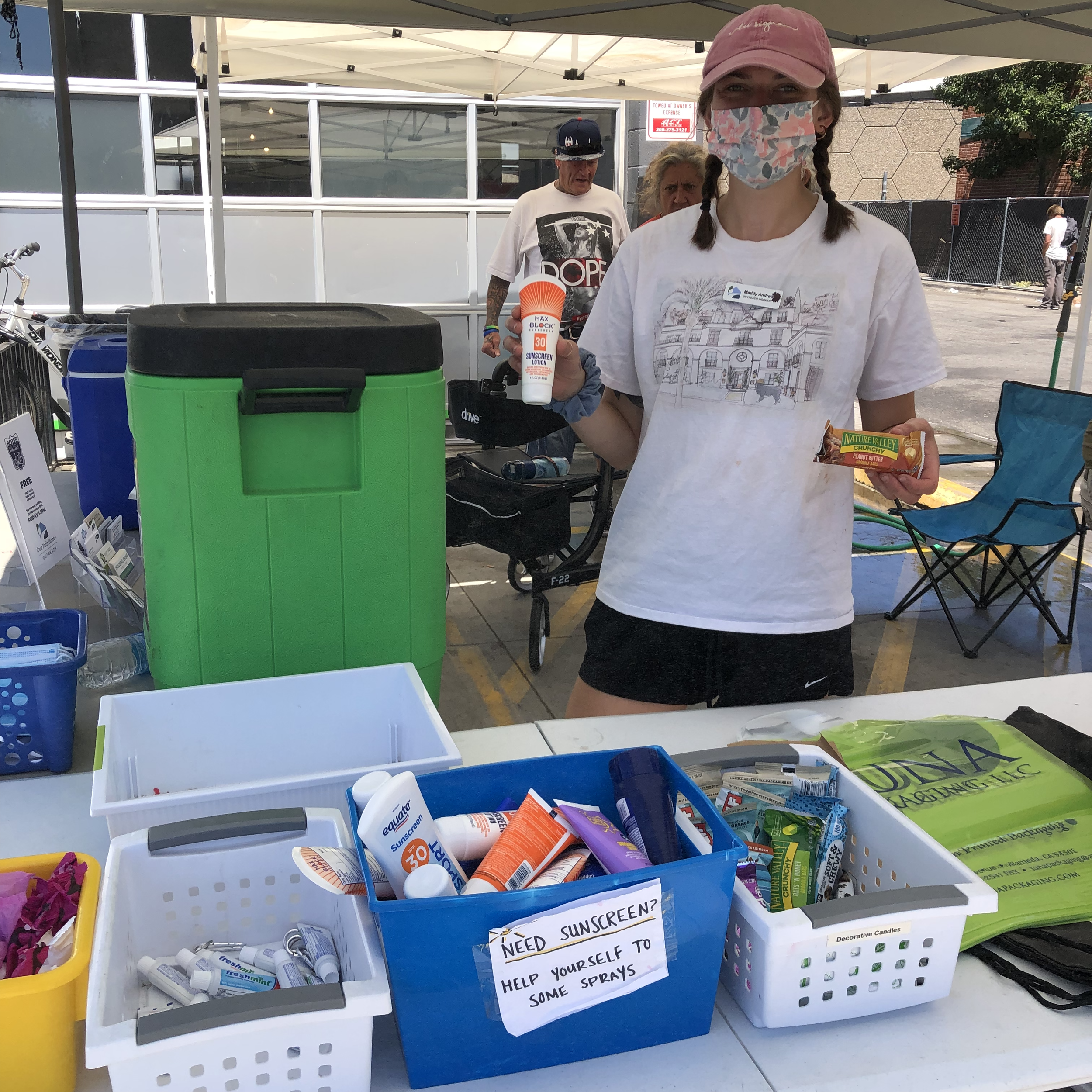 A woman standing behind a display table at an outdoor event, holding a tube of sunscreen lotion and a packet of peanut butter. The table is filled with various items including sunscreen, skincare products, and informational signs about sunscreen needs.