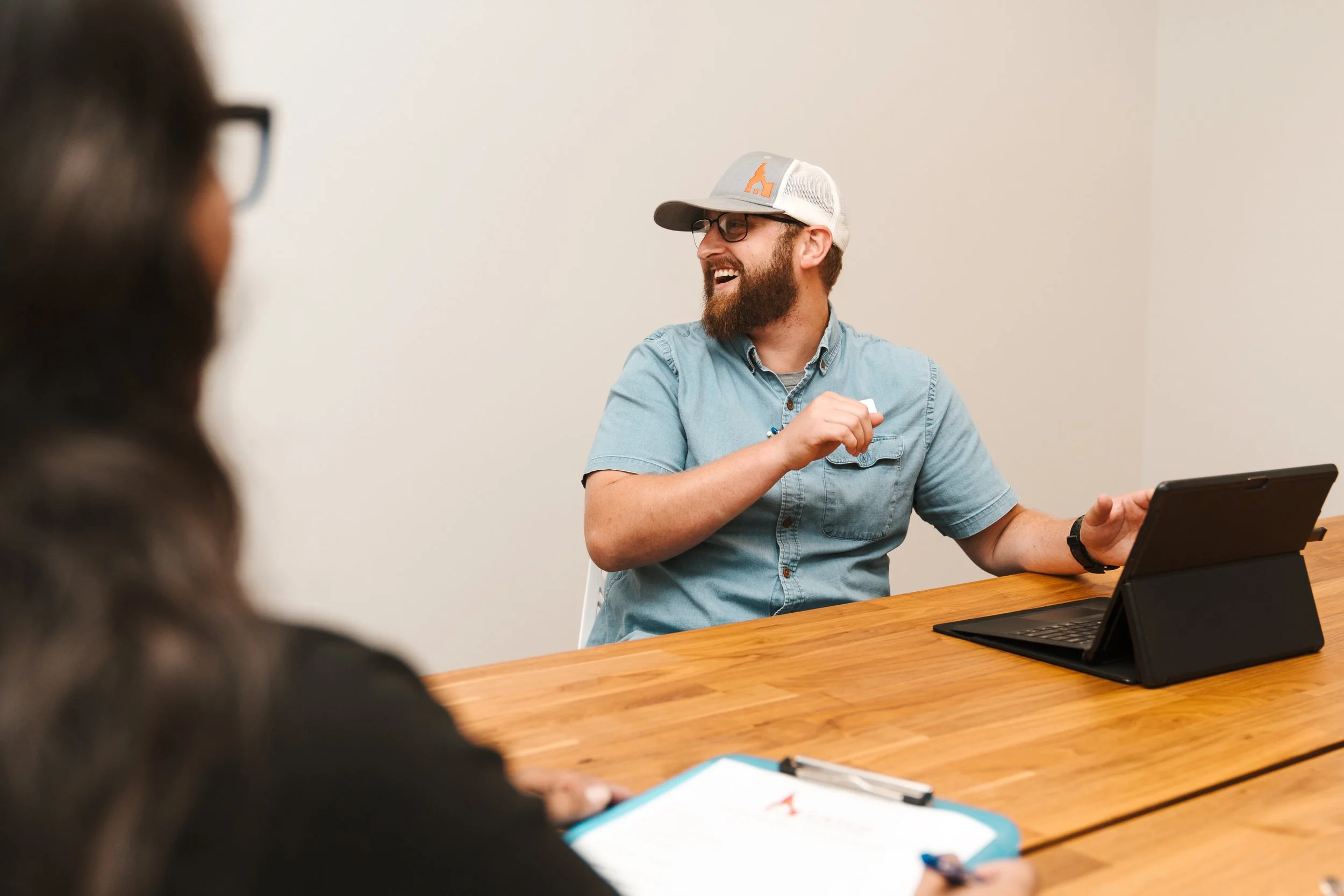 A man with glasses and a beard is smiling and talking, sitting at a wooden table with a laptop and a tablet. A woman with dark hair and glasses is in the foreground, taking notes.