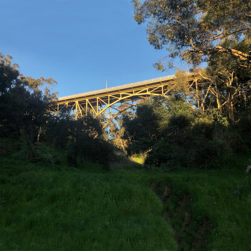A large yellow metal bridge spans above a green landscape with trees and grass, under a clear blue sky.