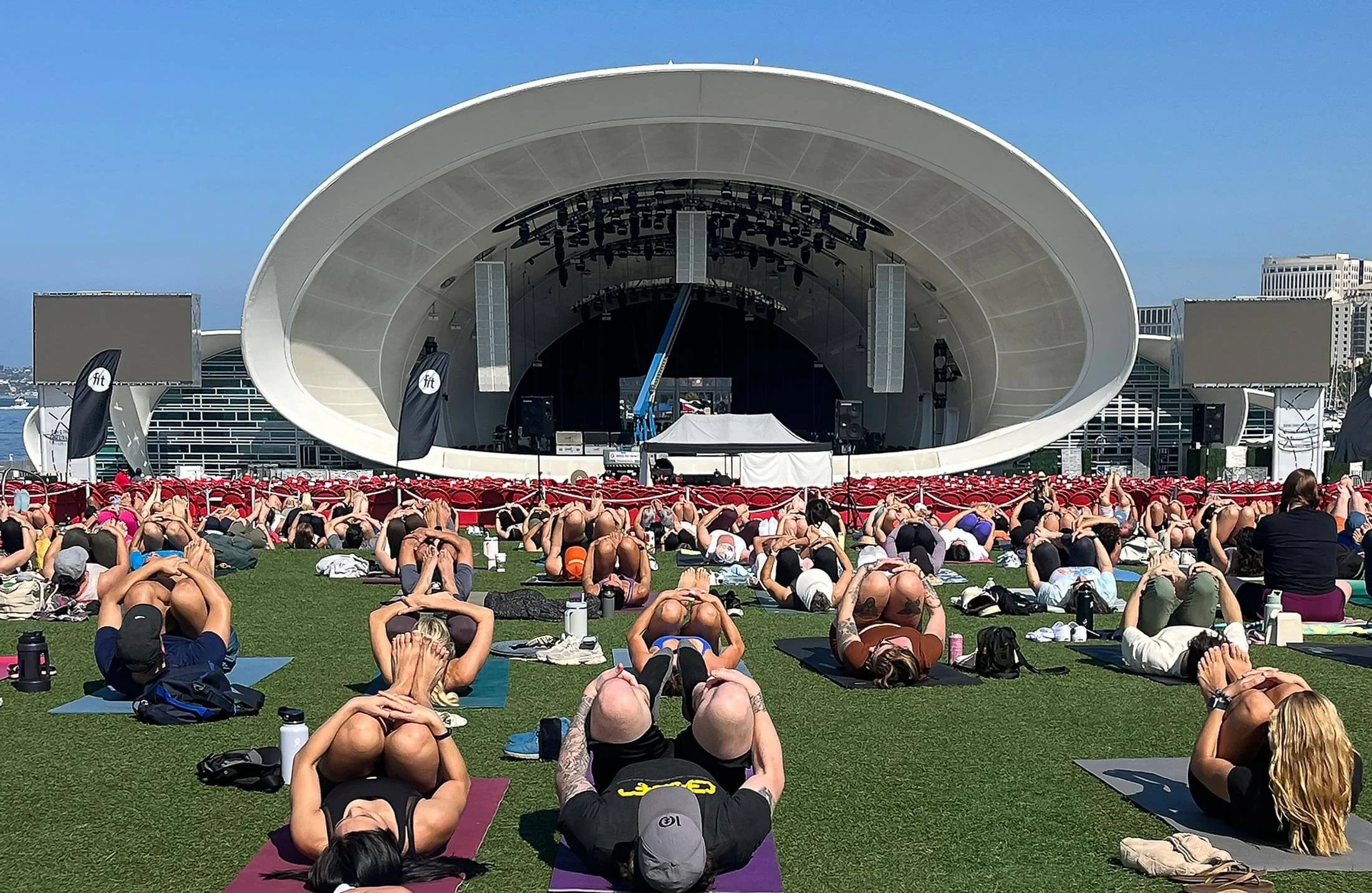 Large outdoor stage with a crowd of people practicing yoga on the grass in front of the stage, under a clear blue sky.