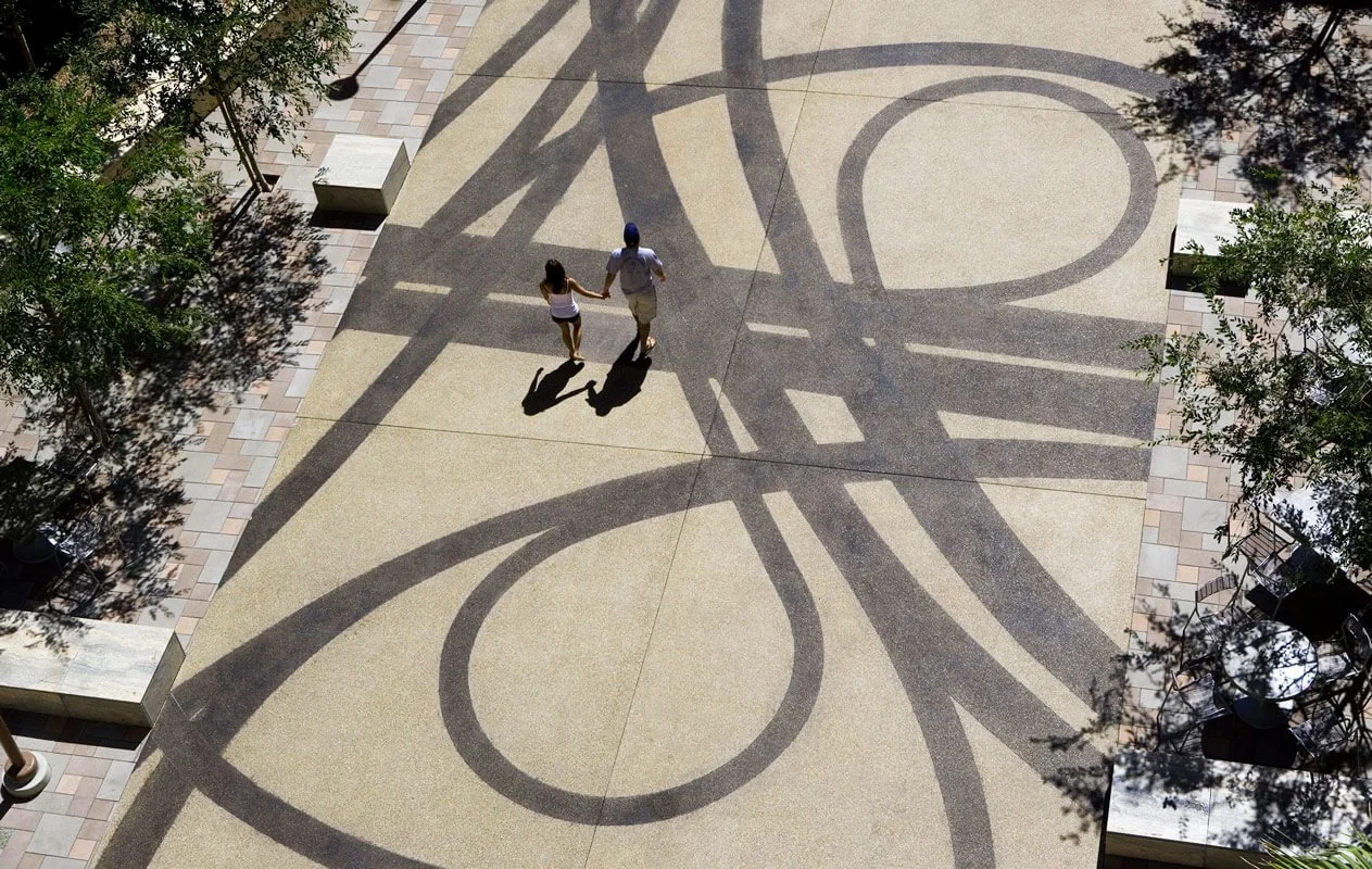 A couple of people walking hand in hand on a paved area with large bicycle shadow patterns cast on the ground near trees and outdoor seating.