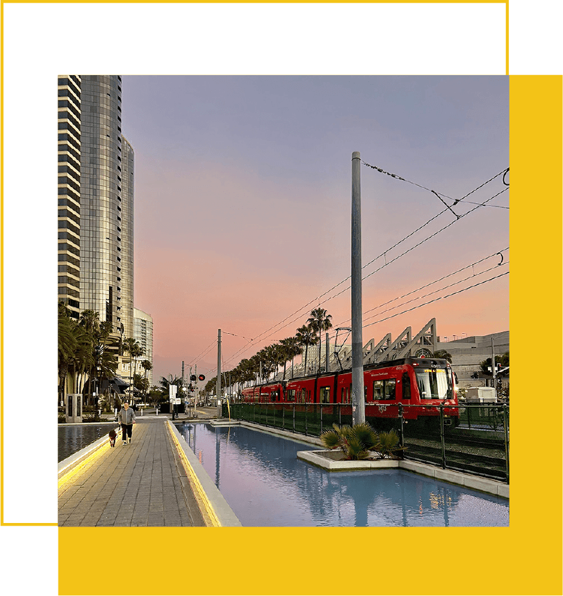 An urban scene at dusk featuring a modern, curved glass skyscraper, a red train on tracks, a palm tree-lined street, and a walkway with people, set against a pastel-colored sky.